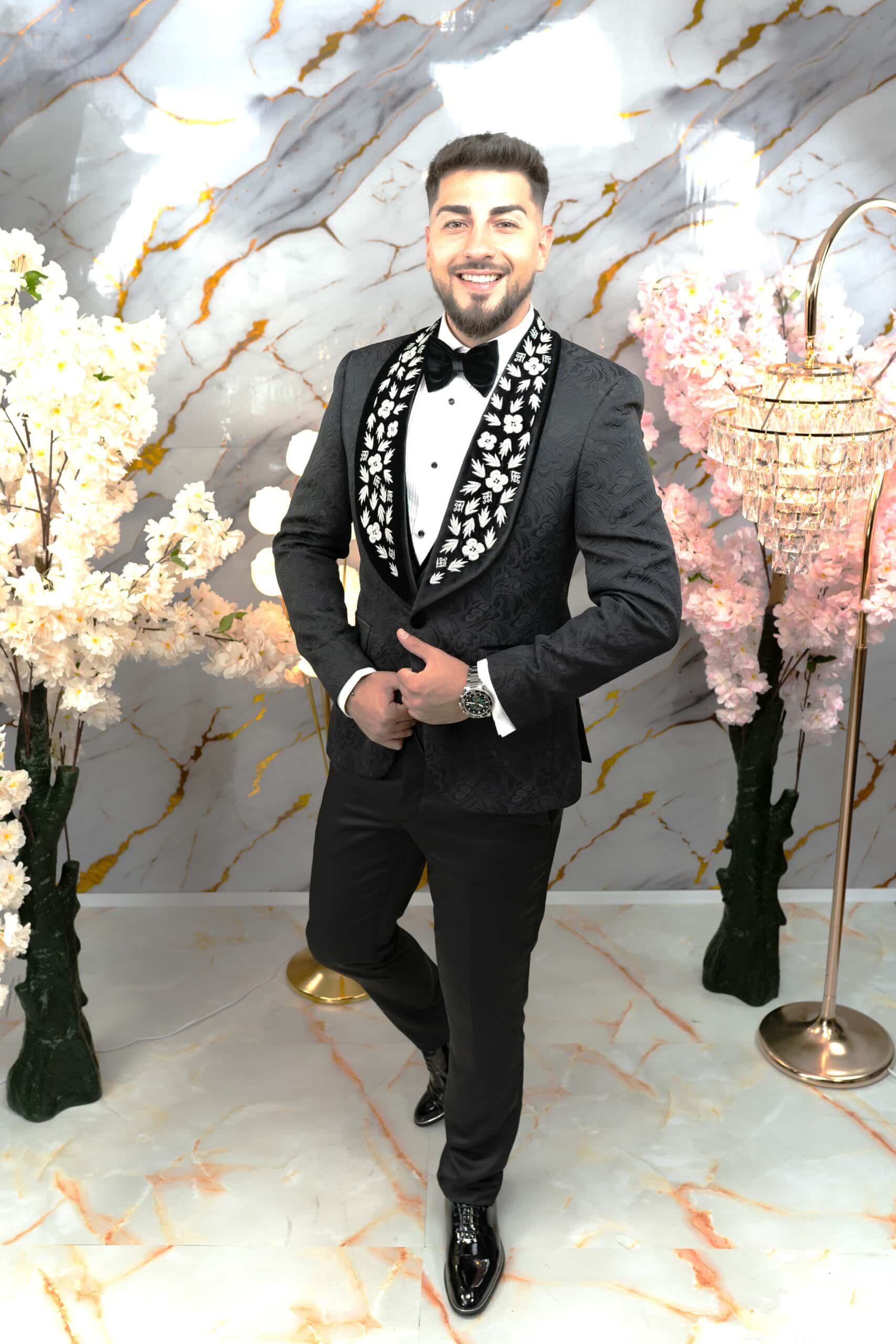 Costume & Pantofi Style Bărbat îmbrăcat într-un costum elegant cu detalii brodate, zâmbind în fața unui fundal decorativ.
